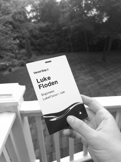 A hand holding the badge to VercelShip 2025.  The background is a large yard with trees further away. Subject appears to be on a balcony. The badge reads "vercel ship, luke floden, engineer, lukefloden.com", with the vercel logo at the bottom right. There is also some fluid with light reflections printed on the bottom third of the badge.