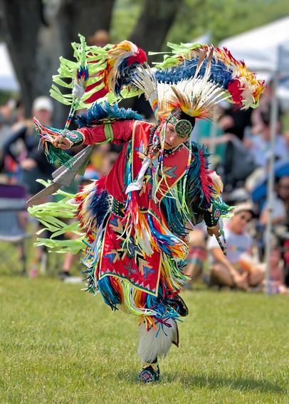 An Indigenous dancer performs at an outdoor powwow, captured mid-dance while balanced on one foot with the other leg raised behind them. They wear vibrant traditional regalia featuring a bright red base garment decorated with geometric patterns in blue, yellow, and turquoise. Large lime-green feather bustles extend from their arms and back like wings. The dancer holds decorated items in both hands and wears a roach headdress with radiating feathers. They perform on grass with seated spectators visible but blurred in the background. The photograph captures dynamic movement and cultural celebration at the 2-Spirit Powwow in Toronto.