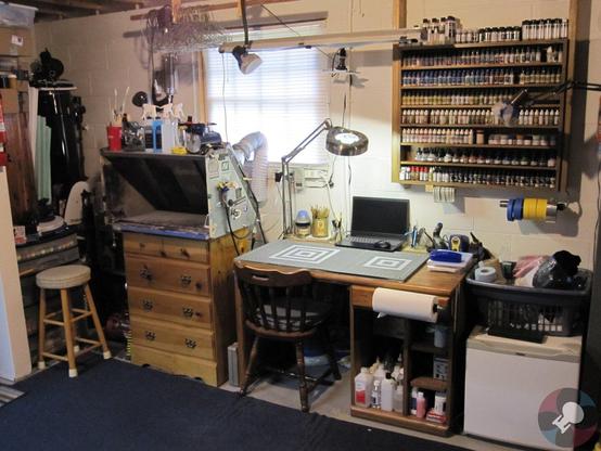 Picture of a hobby workspace for building scale models. A old computer desk with chair in the center and a lit articulating lamp. A spray booth to the left on top of an old kiddy-dresser and stool. A mini-fridge on the floor to the right and hanging paint rack on the right wall. Various tools and supplies are stored about and tucked in various places. Concrete floor with blue carpet and white painted cinder-block wall. A window for booth venting is currently closed and the blinds are down.