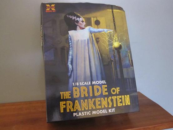 A box of the X-Plus Bride of Frankenstein model kit. On a wooden shelf and gray background. The illustration of the Bride has her arm extended over and electric generator with yellow sparks shooting out. The painted background is of her operating table and the doctor's lab. Main text is in yellow and smaller text in white. The kit is 1/8 in scale.