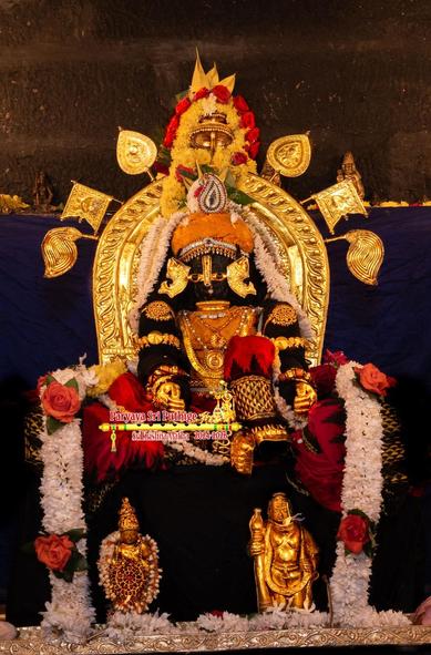 Idol of Hindu deity Krishna in black and red seated with the right leg folded at the knee and lying flat, left leg folded at the knee but vertical, altogether a lounging posture. The black saree drapes over the seat with the red patterns forming a flair all around Krishna and popping on the folded left knee. Krishna wears a vest and turban made of thick Chandana (sandalwood paste), as well as gold shoes, gold palm coverings, several gold ornaments, and a peacock medallion in the turban. 

Krishna leans back onto a gold chairback which itself has six gold flairs, four of those with icons of his usual four weapons: conch, disc, mace, and lotus. A thick dense string of white flowers (with big red roses between) hangs down from the chairback along Krishna's sides and down to an ornate silver slab. Small gold idols on the silver slab, one on either side of Krishna: idol of withala on Krishna's right and a replica of Udupi Krishna on the left.
