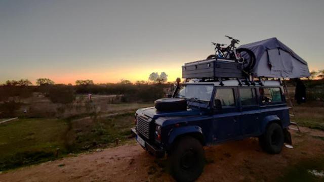 Overland vehicle at sunset in Bubulcus & Bolotas Camping, with a rooftop tent, storage boxes, and bicycles mounted on top. The surrounding landscape is natural and peaceful, with trees silhouetted against a golden sky.