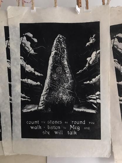 Photo of a number of linocut prints in black ink hanging from a rail. Centrally one print is fully visible. The printed image shows a massive standing stone decorated with circular rock art before a turbulent sky of clouds and force lines. At the foot of the stone is grass and a single foxglove. At the base of the print are the words 'count the stones as 'round you walk listen to Meg and she will talk' in a script-style typeface reminiscent of the gothic work of William Morris. The paper is lightly toned with irregular deckled edges' and is not fully rectangular. At the top of the photo can be seen the peg used to suspend the image on the rail.