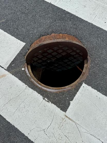 A manhole with its cover half in the hole and half out of it. The manhole is in a pedestrian crossing. 