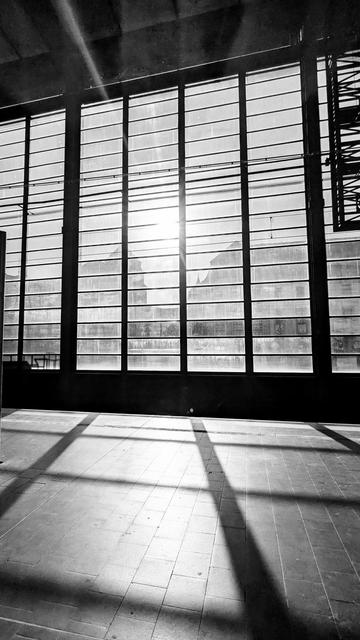 Ein schwarz-weiß Foto.
Blick über einen Bahnsteig auf ein großes Fenster. Dahinter die Sonne und Umrisse von Häusern. Die Streben der Fenster werfen Schatten auf den Bahnsteig.