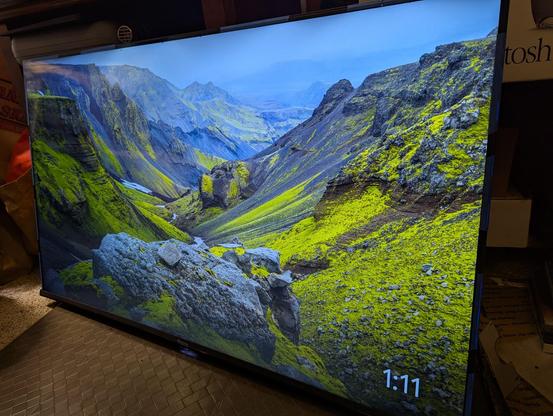 A photograph of a flat panel television, displaying a mountain scene.  A time of 1:11 shows in the bottom right corner.  Strips of blue painter's tape are wrapped around the edges to keep the glass front panel physically connected to the back panel.