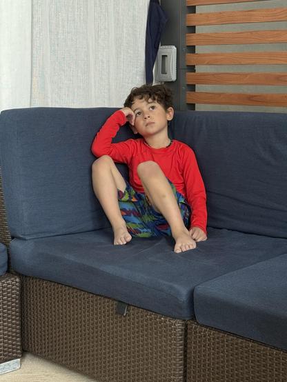 A young child sits on a blue couch, resting his chin on his hand, appearing contemplative or bored. He wears a red long-sleeve shirt and colorful patterned shorts, with bare feet visible. The background includes light-colored curtains and wooden sl