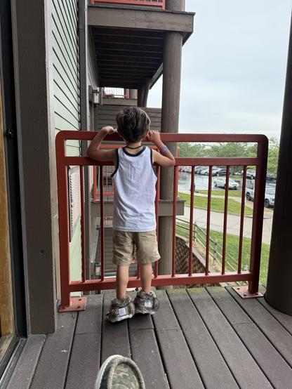A child is standing on a balcony, leaning against a red railing. The child is wearing a white tank top, beige shorts, and fuzzy slippers. The background shows a view of a parking lot and greenery, with cloudy weather overhead.