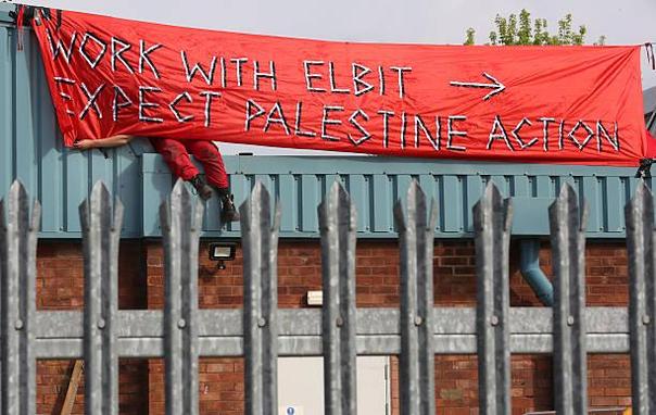 Banner text:
W O R K    W I T H    E L B I T
E X P E C T    P A L E S T I N E    A C T I O N

Photograph through metal spike fence shows a red banner and a person’s legs 🦵 in red overalls seen beneath the banner over the edge of the building roof. 