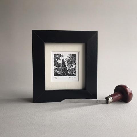 Photo of a dark, square picture frame on a table, with a wood engraving tool before it. In the frame is a black ink printed image of a standing stone with circular decorations under a dramatic, swirling sky