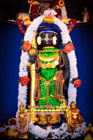 Idol of Hindu deity Krishna in bright green standing with a gold-covered staff in right hand; wearing gold shoes, a blue silk turban, and a broad gold necklace with many coin-like medallions hanging from the necklace. A thick dense string of white flowers (with big red roses between) all around Krishna. Small gold idols on the silver slab, one on either side of Krishna: withala on Krishna's right and a replica of Udupi Krishna on the left. Blue cloth background.