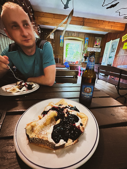 Wnętrze schroniska. Kadr przy stole. Na przeciwko człowiek trzymający widelec, za pomoc którego wcina naleśniki z jagodami. Na pierwszym planie talerz z takimi samymi naleśnikami. Obok stoi butelka czeskiego piwa bezalkoholowego. 