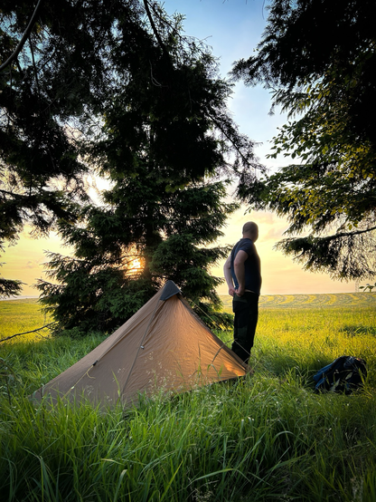 Rozbity namiot, obok postać na tle zachodzącego słońca. Dookoła świerki. W oddali skoszona łąka. 