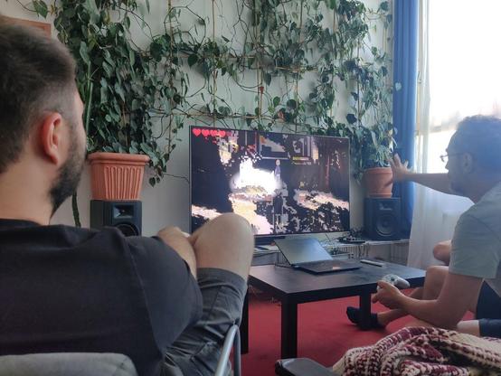 Team ROTxBLAU sitzt auf Couch und Sesse, vor einem Bildschirm, auf dem eine Version von Meine Oma (88) läuft. Auf dem Bildschirm ist zu sehen, wie die Welt, die die Spieler:innen sehen, zerbricht und glitcht.