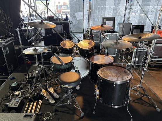 Drum kit on the side of the stage a few minutes before playing a gig. The drum kit has 4 toms, a snare drum, bass drum with double bass pedal, hi-hat, 2 crash cymbals, a ride and a China cymbal. On the floor, an audio interface, several pairs of drum sticks. A tablet is mounted on one of the cymbal stands.