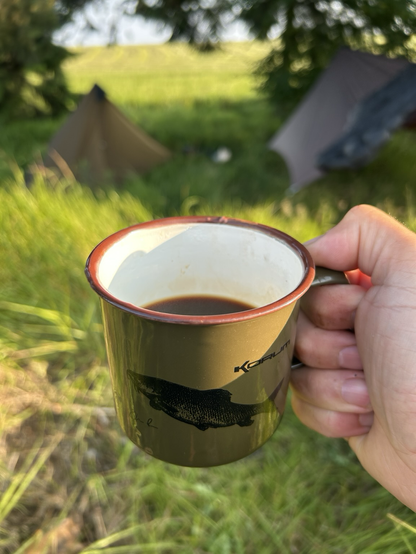 Dłoń trzymająca emaliowany kubek z kawą. W tle rozbity namiot oraz rozwieszony pomiędzy drzewami hamak. 