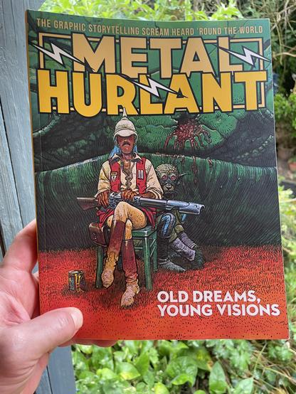 A white male hand holding up a copy of 'Metal Hurlant: Old Dreams, Young Visions' in front of some wet foliage.