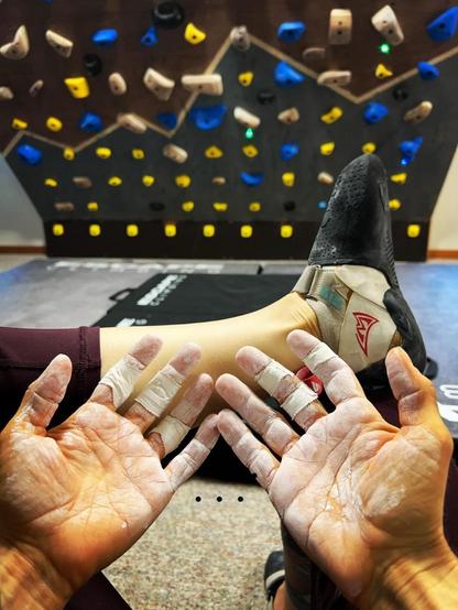 My open rough hands covered in chalk and taped where the skin has torn from climbing. In the background you can see a Moonboard (an angled climbing board with holds).