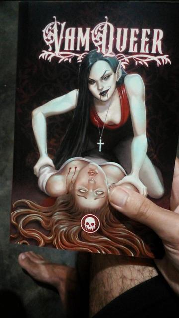 Minha mão segurando o livro. Descrição da capa: Uma vampira sensual de blusa vermelha sentada sobre uma mulher deitada. A vampira tem orelhas pontudas, cabelos lisos pretos e maquiagem escura, com duas listras de sangue escorrendo dos lábios e um colar com crucifixo pendendo sobre os seios. Ela prende a mulher no chão, segurando seu ombro. A mulher, com os cabelos loiros se espalhando no chão, olha para nós com rosto aflito, como se pedisse socorro. Tem os olhos sem pupila e duas marcas ensanguentadas de caninos sobre o seio.