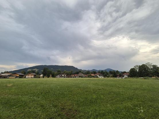 A serene and picturesque scene unfolds in this image, showcasing a large grassy field stretching out under a clear blue sky. In the distance, a cluster of houses can be seen nestled amongst the lush greenery. A solitary tree stands tall in the foreground, adding a touch of natural beauty to the landscape. The atmosphere is peaceful and inviting, with fluffy white clouds drifting lazily overhead. This idyllic rural setting is the epitome of tranquility, offering a glimpse into the harmony of nat…