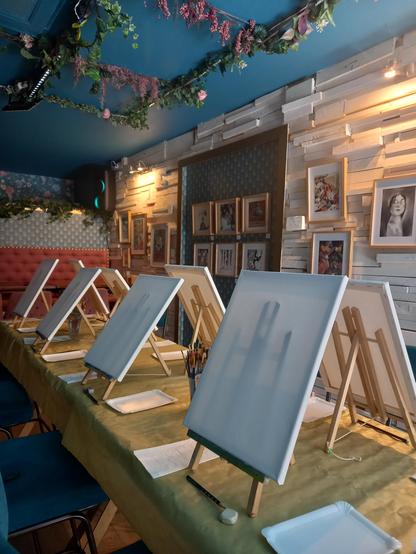 Foto al interior del Amarone Vinneria. Se ve en diagonal una mesa larga preparadas con mantel dorado, caballetes con lienzos en blanco y paletas. Detrás, en la parte de la derecha, se observa la exposición temporal de ilustraciones de Toño Benavides.