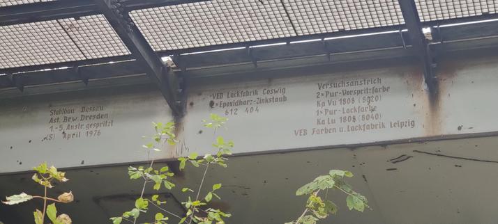 Auf dem Bild ist ein Teil einer Eisenbahnbrücke aus Stahl zu erkennen.
Diese ist mit einer Grau-Grünen Rostschutzlackierung versehen. Es handelt sich dabei um eine Versuchslackierung wie man den Text der sich auf Ihr befindet entnehmen kann:

Stahlbau Dessau
Außenstelle Brw Dresden
1. - 5. Anstrich gespritzt
(S) April 1976

VEB Lackfabrik Coswig
2(?)x Epoxidharz-Zinkstaub
 612 404

Versuchsanstrich
2 x Pur-Vorspritzfarbe
Ka Vu 1808 (8020)
1 x Pur-Lackfarbe
Ka Lu 1808 (8040)
VEB Farben und Lackfabrik Leipzig
