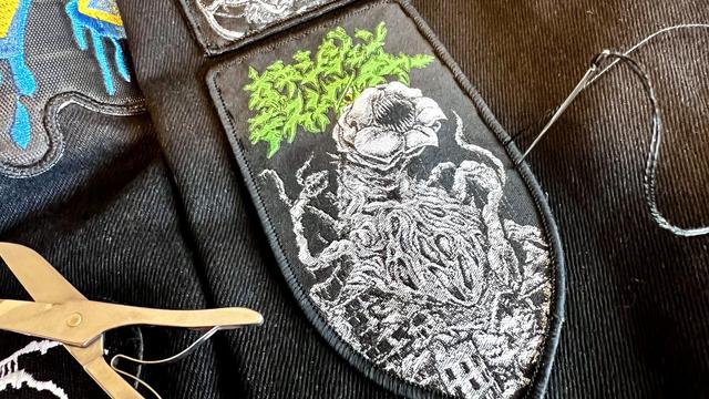 A black, shield shaped, Oxygen Destroyer patch halfway sewn on the right arm of a black denim battle jacket. Scissors from a Swiss Army knife can be seen peeking from the left side of the frame. 