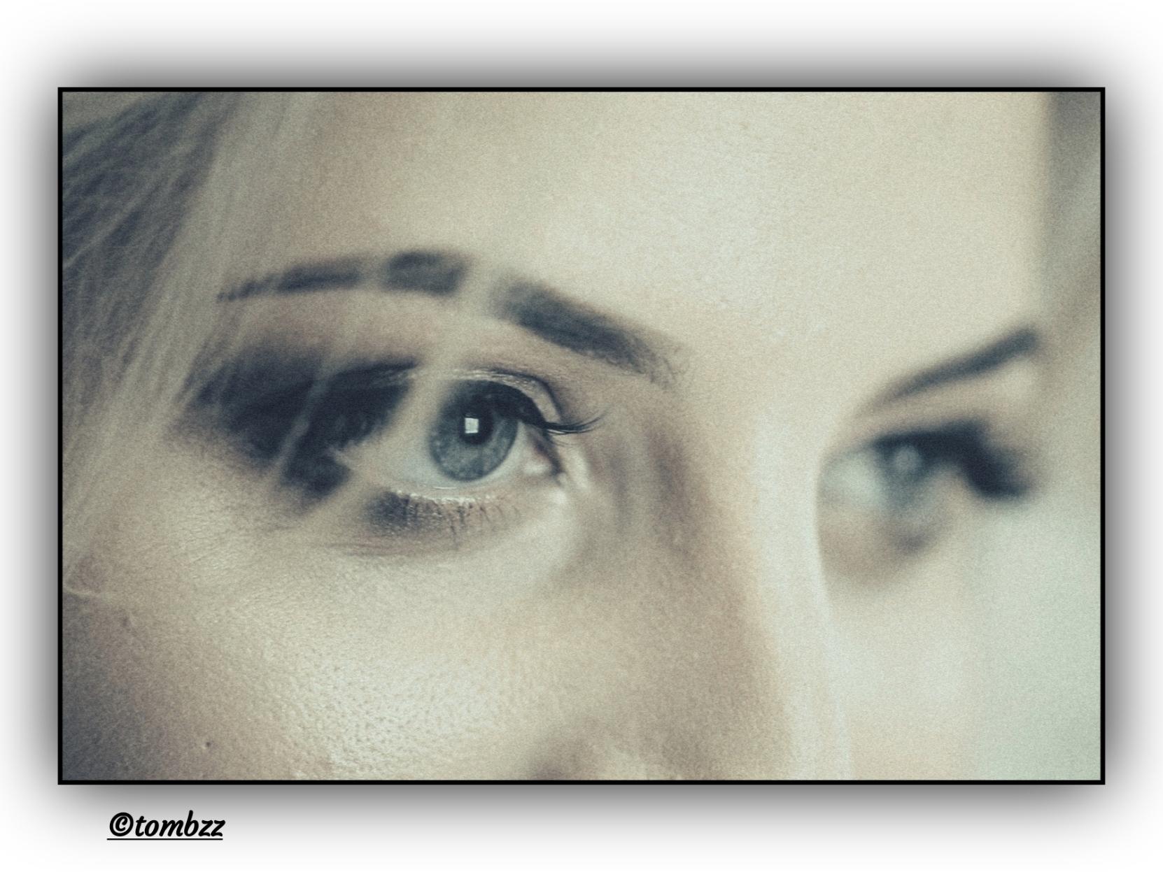 Close-up of a young woman's face captured on film. Her eyes are light blue, full of depth and introspection. The gaze — intense, thoughtful, as if a moment of inner dialogue has been caught. Thick, long eyelashes subtly emphasize the shape of her eyes. Her brows, dark and naturally shaped, add character. The soft grain and lighting typical of analog photography give the image a nostalgic, gentle quality.