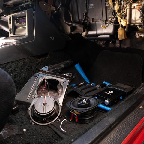 A car's right-side footwell with the dashboard partially disassembled, revealing speaker wiring and a scattered toolkit. An iFixit soldering toolkit sits in the footwell.
