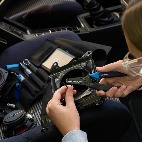 Close-up of someone using iFixit wire strippers to strip the tips of speaker wires inside a car. The work mat is loaded with tools, speakers, and parts.