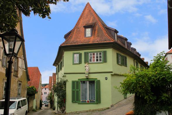 Blick auf ein Eckhaus an einer abschüssigen Straße (eher Gasse). Das Gebäude ist hellgrün gestrichen und hat grasgrüne Fensterläden. Das Dach ist mit inzwischen verwitterten Ziegeln gedeckt. 