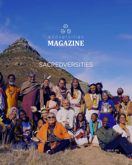 A diverse group of smiling people wearing a variety of ethnic and indigenous clothing standing and sitting on the sandy ground in front of a scraggly hill and blue skies.