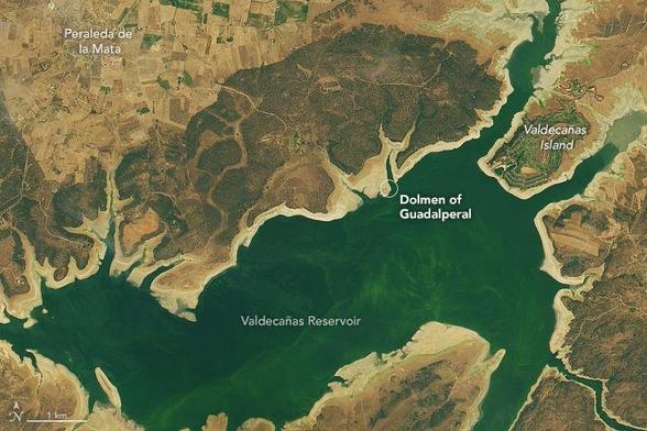 This is a satellite photograph centered on Valdecañas Reservoir in western Spain. A circle on the northern coastline is labeled "Dolemen of Guadalperal" and is now mainly on the shore. The city of Peraleda de la Mata is in the upper left, and Valdecañas Island is in the upper right.