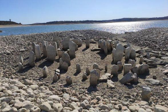 This is a close-up photograph of a set of standing stones on the shoreline of a body of water, standing in rings and surrounded by smaller rocks outward to the water. The sky above is blue and cloudless, and a far shoreline can be seen in the distance.