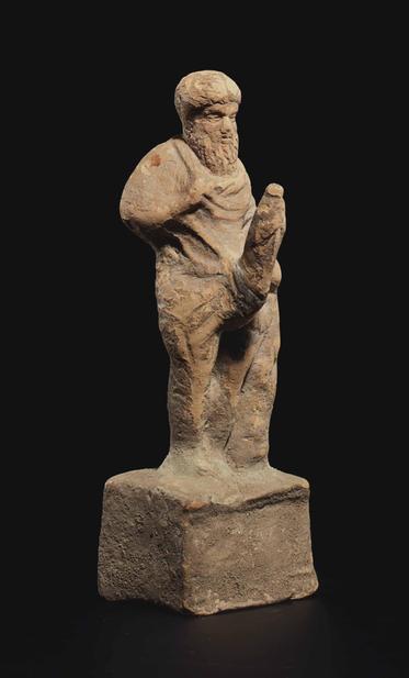 Photograph of a Greek terracotta Priapos against a black background. The fertility god stands with his feet together on a high integral plinth, leaning against a tree trunk to his left, his left hand resting on top, depicted with a full beard, his long center-parted hair pulled back and gathered in a chignon at the base of his neck. His body is enveloped in a himation but his erect phallus is fully exposed, even the glans, which was considered obscene in ancient Greece.