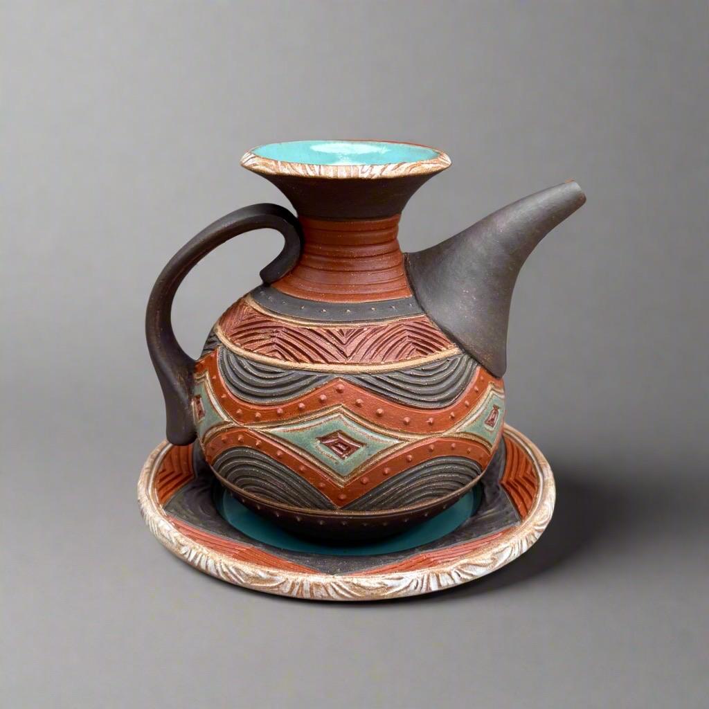A decorative ceramic ewer with a spout, featuring intricate geometric patterns and vibrant colors, accompanied by a matching plate. The surface is decorated with carved lines and small raised dots. A row of aqua diamonds encircles the rotund pot like a belt through the center. The jug has a curved handle and a wide, flared opening. The colors are black and dark red with deep aqua accents.