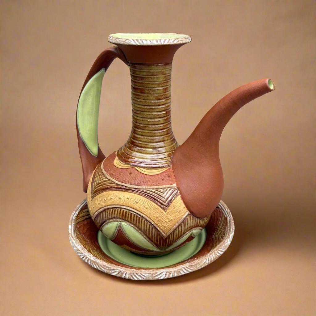 A decorative ceramic ewer with a long spout and an elongated handle, featuring vibrant colors of dark orange, mottled brown-green, dark yellow with bright chartreuse accents. and geometric patterns. The long neck features concentric rings and the round body of the ewer is decorated with geometric patterns, carved lines and tiny raised dots. It rests on a shallow, matching plate. The background is a warm tan color.