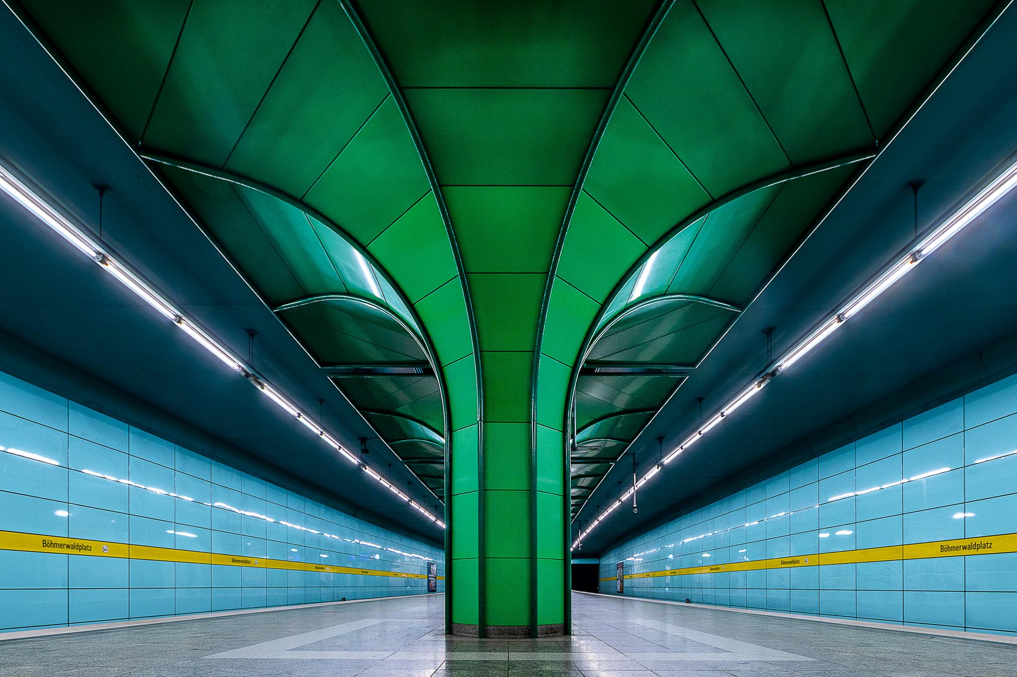 Symmetrical view of Böhmerwaldplatz metro station in Munich with an impressive green pillar spreading on the ceiling like a tree in the foreground, in the middle of the platform. Symmetrical view of Böhmerwaldplatz metro station in Munich with an impressive green pillar spreading on the ceiling like a tree in the foreground, in the middle of the platform.