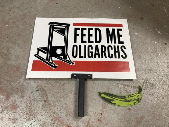 A sign with a guillotine that says "Feed Me Oligarchs" with a laser-cut banana next to it.