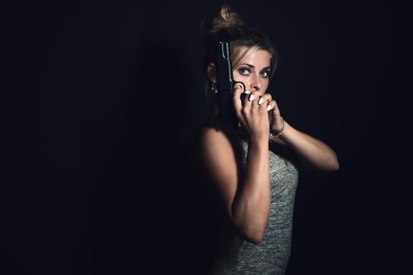 An attractive woman holding a pistol in both hands while posing for a side profile in dark light.