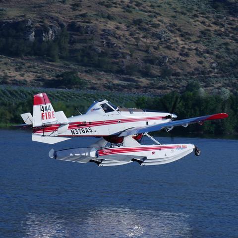 Another photo showing the same Air Tractor AT-802 FireBoss aircraft from it's right side. It has a red tipped vertical stabilizer, a multi-lined expanding red stripe on the side, and a similar stripe on the floats. The wing tips are red. It has a single person cockpit in the center of the aircraft that has a trailing "right" flag behind the right window.