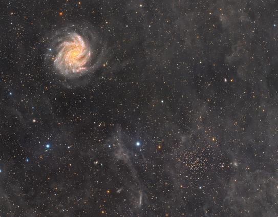 A wide-field astrophotograph showing two main celestial objects. In the lower right is NGC 6939, a tight grouping of bluish stars forming an open star cluster. In the upper left is NGC 6946, a bright face-on spiral galaxy with dusty arms, tinged in subtle red and blue hues. Faint interstellar dust clouds stretch across the image, giving it a textured appearance. The entire scene is filled with tiny stars from our Milky Way in the background.