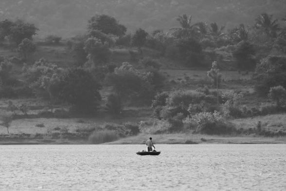 A fisherman standing on a coracle in a lake against the backdrop of hills and vegetation in the background. The photo is in black and white.