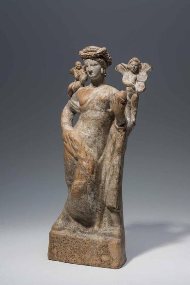Terracotta figurine of Aphrodite, fully dressed, leaning on a plinth or tree. She rests her right hand on her hip, holding an apple or a flower with her left. Two winged Erotes are with her, one standing on the plinth or tree, the other sitting on her shoulder.