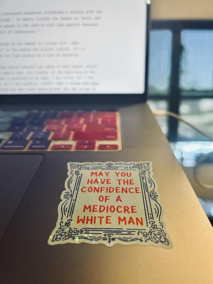 A sticker with the text 'May you have the confidence of a mediocre white man' affixed to my laptop next to my rack pad. The essay I'm working on is visible yet out of focus on the computer screen.