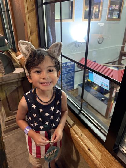A young boy with brown hair is wearing a tank top featuring an American flag design and a headband with fluffy ears. He is smiling and holding a badge labeled "PUP" while standing near a window with a view of a brightly lit space.