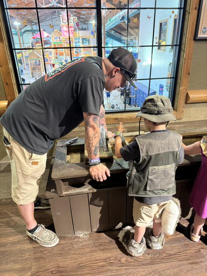 A man and a child are leaning over a wooden table, engaged in an activity. The man has tattoos and is wearing a black hat and a t-shirt, while the child, dressed in a vest and unique fuzzy footwear, appears to be focused on gem mining 