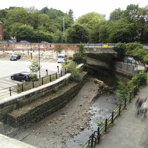 Photograph of the tidal stretch of Ouseburn River at south side of Ouseburn Bridge in Newcastle upon Tyne. To the left (south-west) of the bridge is Cut Bank, and to the right (north-east) is Byker Bank. In the foreground is the pedestrian Riverside Walk and the photo also spans some surrounding built-up areas and trees, with sky in the background. Still image captured from webcam at 17:36 hrs on Friday 4 July 2025.