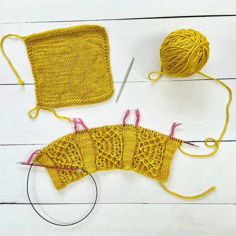 Flat-lay view of the start of a bright yellow knitting project with textured bands of patterning on a circular needle, the ball of yarn, and the swatch on a white plank background.