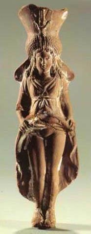 Isis-Aphrodite figurine, probably made from clay. She wears an elaborate headdress and her hair in multiple braids. Her dress girded below the breasts. She lifts her dress's skirt, exposing her vulva.